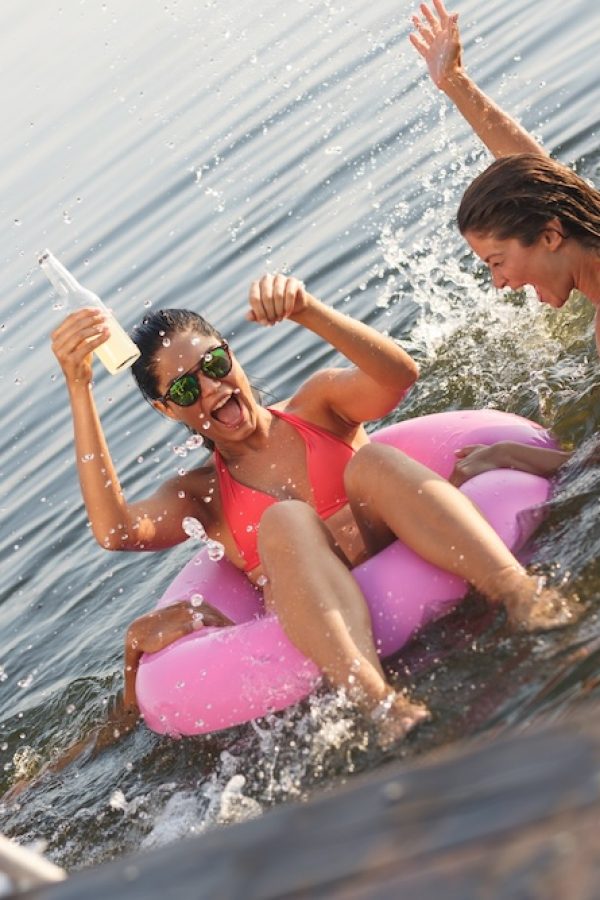 Group of friends swimming and having fun in the lake.Female sitting on air mattress drinks lemonade and having fun with friends.